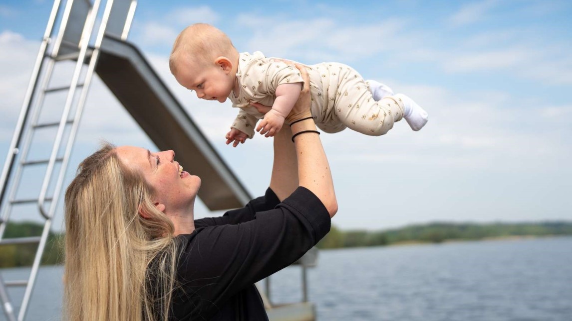 Billede af mor der løfter sit barn højt op mens hun smiler og står foran en sø med en vandrutsjebane